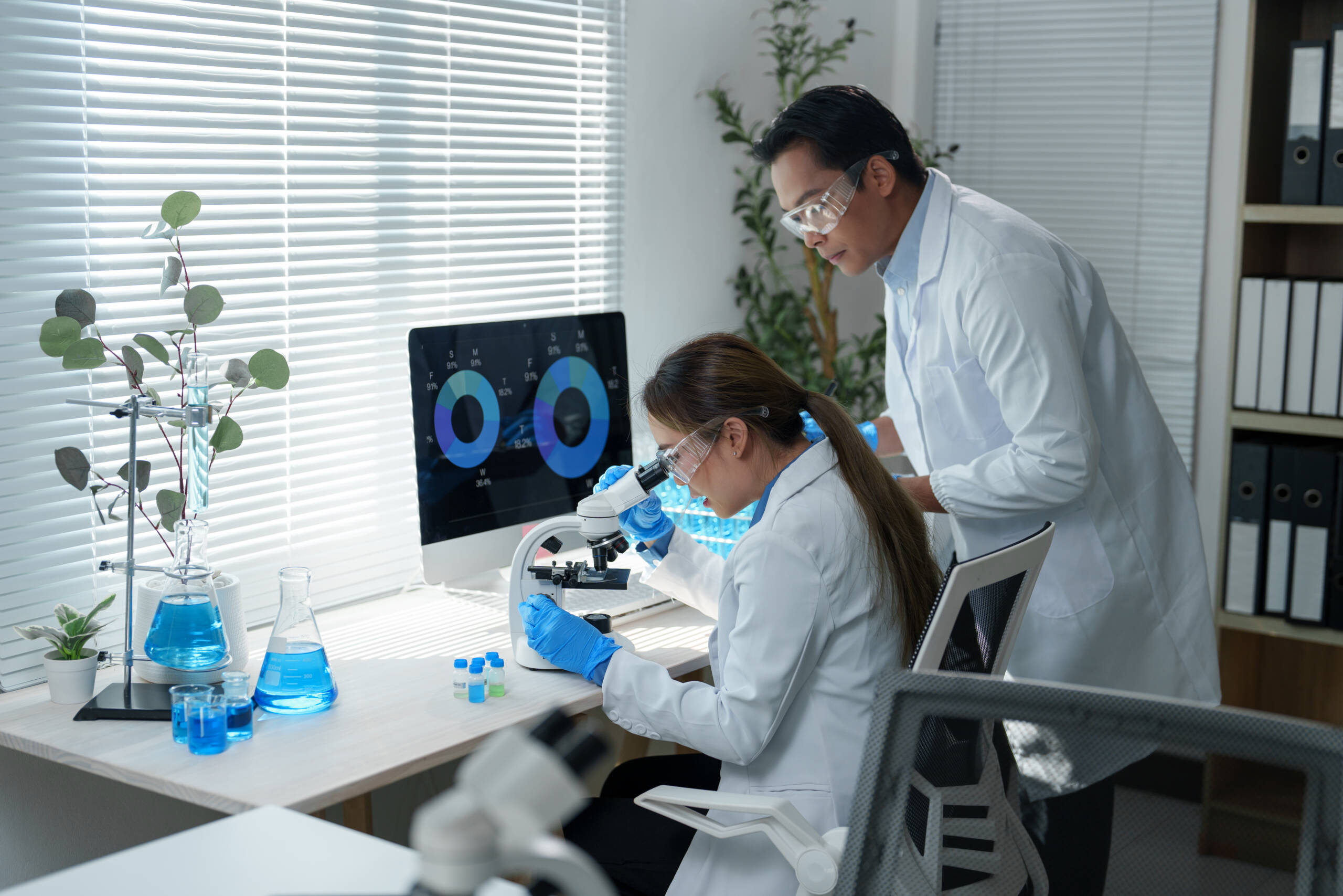 Scientists wearing lab coats and protective eyewear work in a laboratory. One uses a microscope while the other observes data on a computer screen, surrounded by scientific equipment Scientists wearing lab coats and protective eyewear work in a laboratory. One uses a microscope while the other observes data on a computer screen, surrounded by scientific equipment